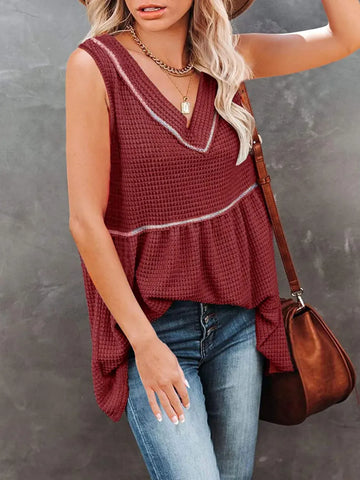 A woman wearing a sleeveless waffle-knit peplum top in burgundy with white contrast trim, paired with blue jeans and a brown shoulder bag, standing against a textured gray wall.