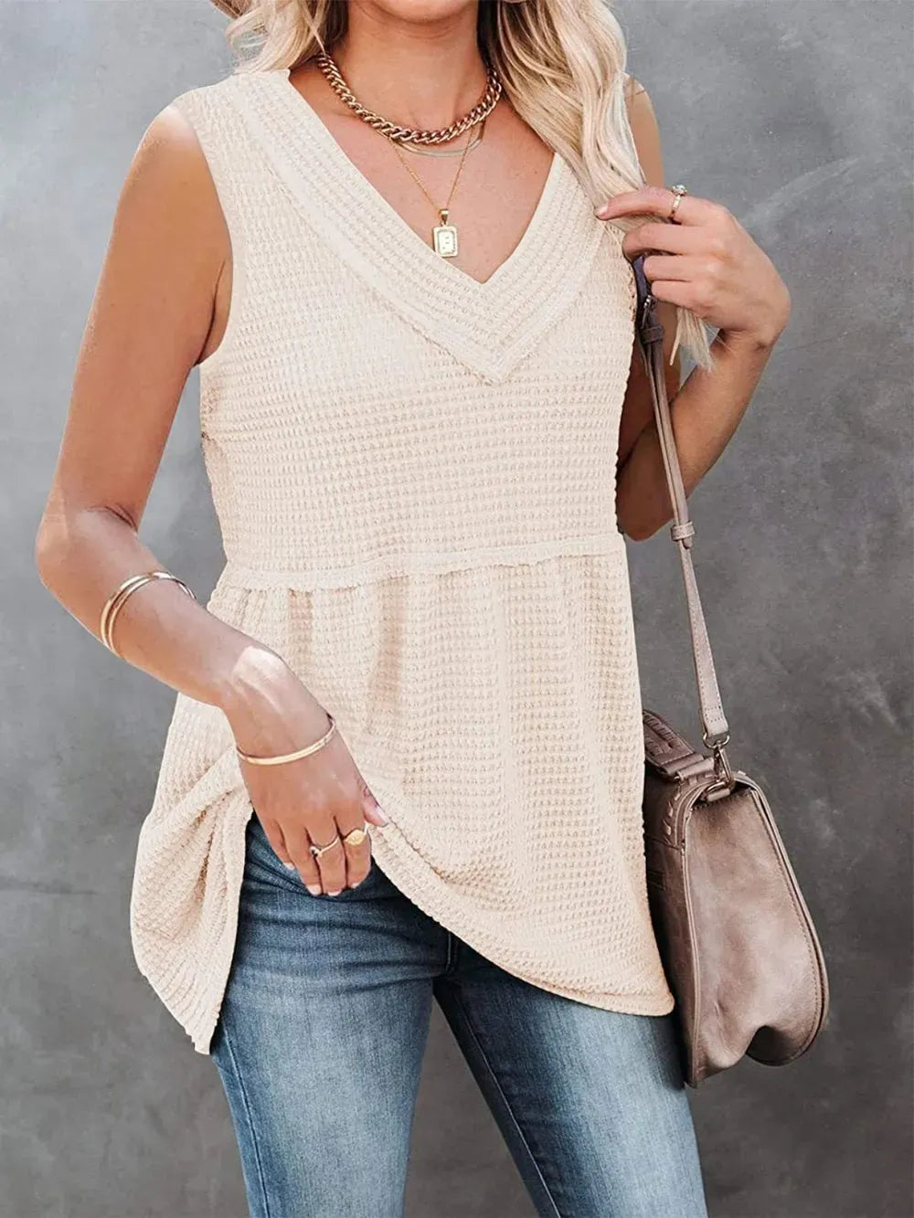 Woman wearing a beige sleeveless top and blue jeans, holding a brown handbag against a gray background