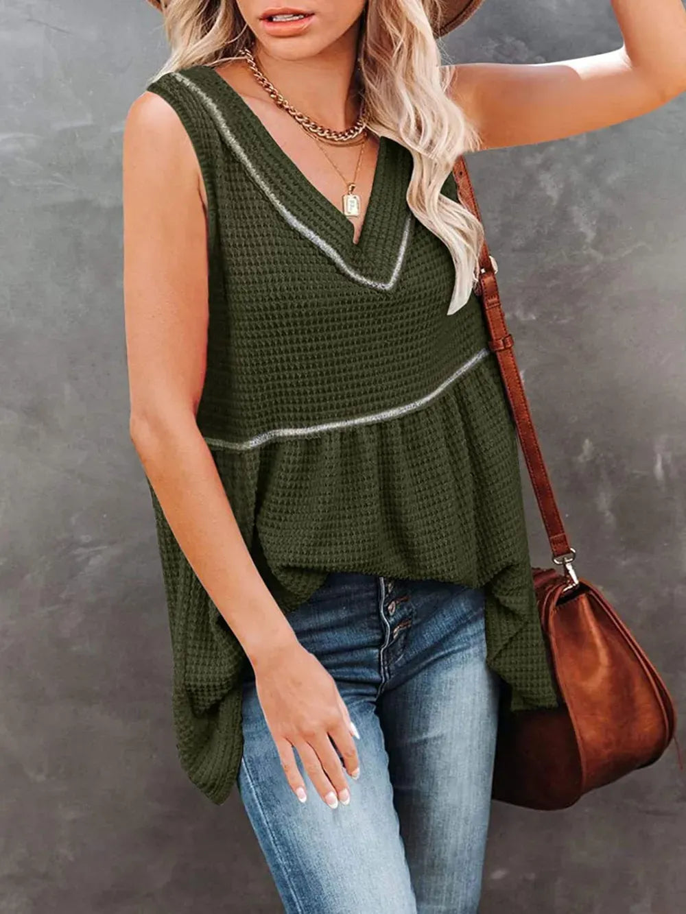 A woman wearing an olive green waffle-knit sleeveless top with a V-neck and peplum hem, paired with blue jeans and a brown shoulder bag, set against a textured gray background.