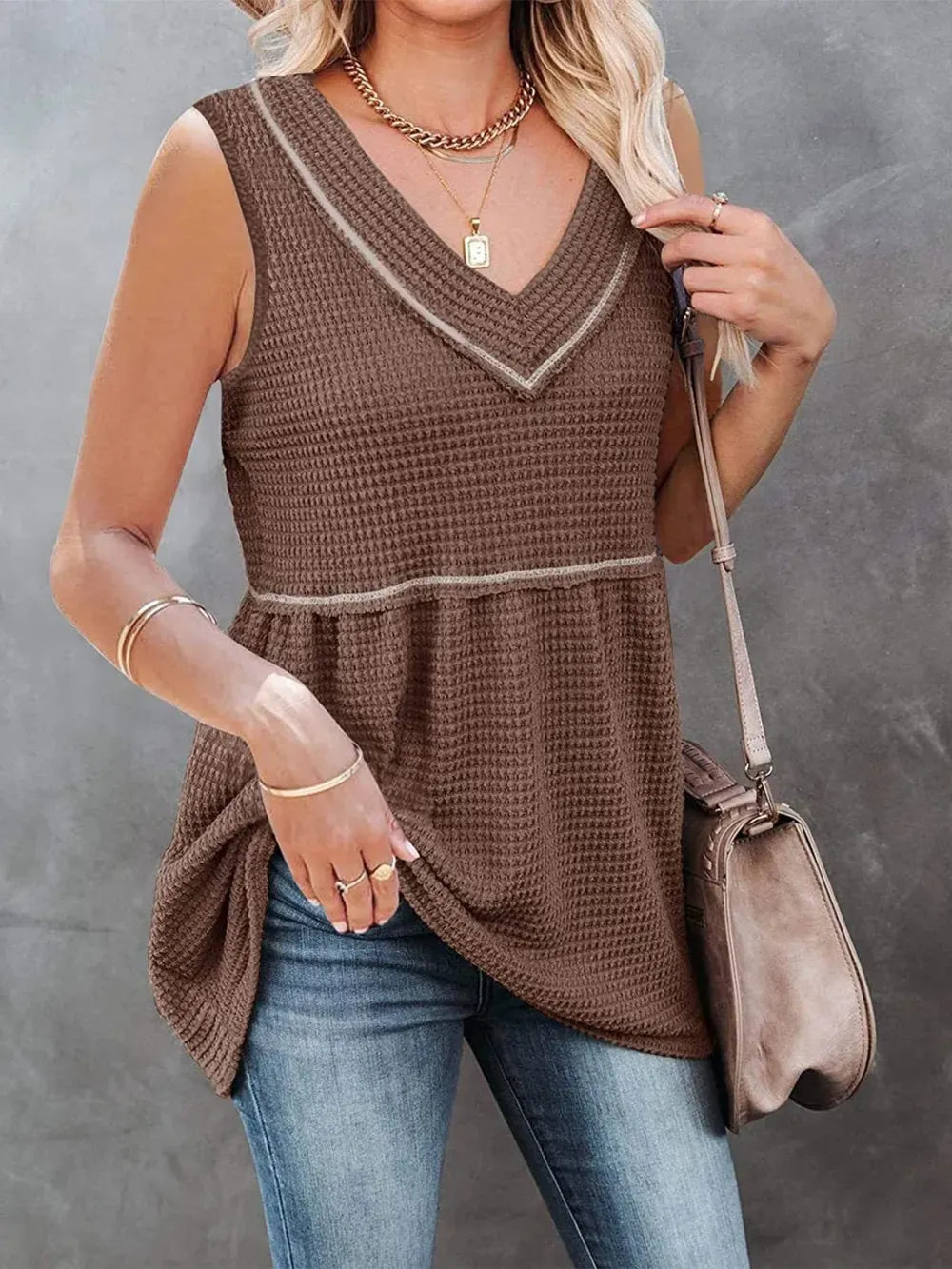 Woman wearing a brown sleeveless top and blue jeans, holding a handbag against a gray background