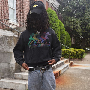 A person standing on outdoor stone steps in front of a brick building, wearing a black sweatshirt with a colorful retro graphic on the front, a black cap, and light blue jeans.