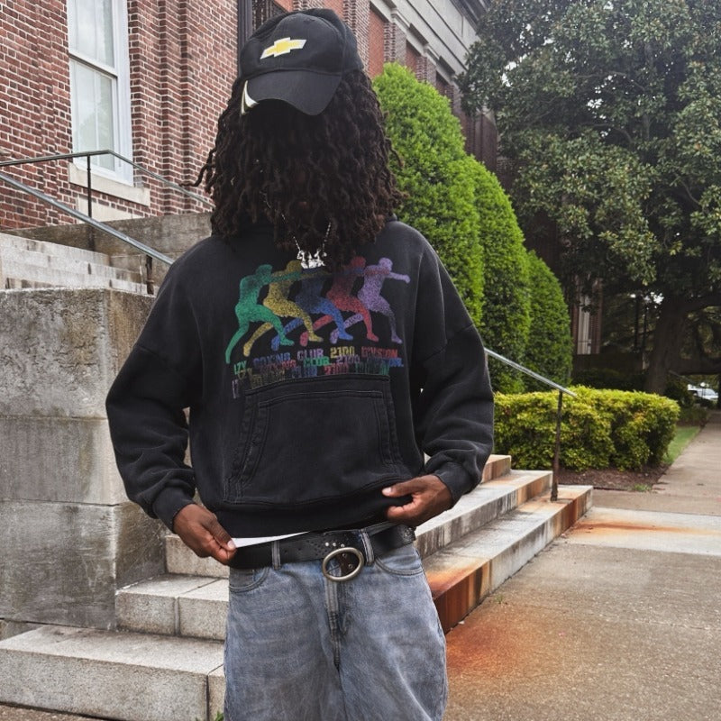 A person standing on outdoor stone steps in front of a brick building, wearing a black sweatshirt with a colorful retro graphic on the front, a black cap, and light blue jeans.